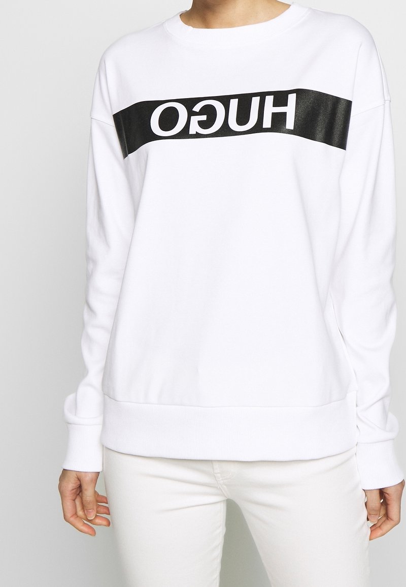 Bílá mikina s černým logem "HUGO" přes přední část. Má kulatý výstřih, žebrované manžety a lem. Měkká textura, pohodlný design.
