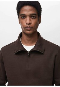 Pullover in pile marrone con collare a zip, caratterizzato da una superficie testurizzata e una maglietta bianca sottostante. Vestibilità su misura con dettagli di stile minimi.