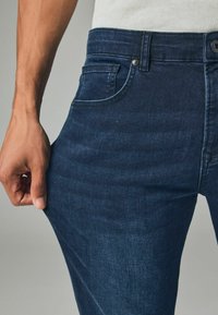 Tmavomodré džínsy s úzkym strihom. Majú jemne vyblednuté časti, päť vreciek a kovové zapínanie na gombík. Ruka drží látku.