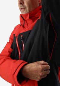 Veste rouge et noire avec des zips, manches ajustables et doublure en maille. Le matériau semble résistant à l'eau ; dispose d'un col montant pour la chaleur.