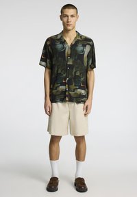 Kortärmad svart skjorta med abstrakta gröna och orange mönster, knäppdesign, ihopparad med beige shorts och bruna slip-on skor.