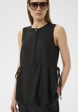 Naine kannab üle suurte pruunide päikeseprillide ja varrukateta musta topi külgede sidemetega ja esiküljel nööpidega detailiga.