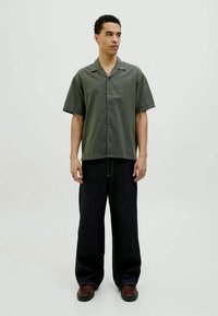 Joven de pie, vistiendo una camisa de botón de manga corta de color verde oscuro, pantalones negros holgados y zapatos marrones, contra un fondo claro y liso.