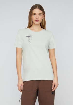 Femme portant un t-shirt gris clair avec un petit motif d'arbre et le texte "MAMMUT", associé à un pantalon marron, debout devant un fond uni.
