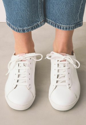 Nohy v bielych teniskách so šnurovaním a krátkych modrých džínsach stojace na hladkej svetlosivej podlahe.