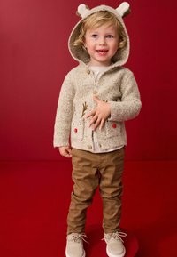 Tan gebreide cardigan met berenoren, geborduurde rendierzakken, knoopsluiting. Gepaard met bruine broeken en lichtgrijze schoenen.