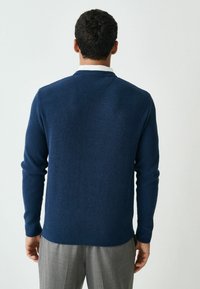 Marineblauer gerippt gestrickter Pullover mit langen Ärmeln und rundem Halsausschnitt, von hinten gezeigt, kombiniert mit hellgrauen Hosen.