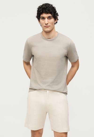 Νεαρός με σγουρά μαλλιά που φοράει μπεζ κοντομάνικη μπλούζα και ανοιχτόχρωμα σορτς, στέκεται με τα χέρια πίσω από την πλάτη.