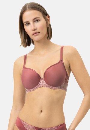Frau trägt mauve gepolsterten BH und passende Unterwäsche mit Spitzenbesatz, steht vor schlichtem hellem Hintergrund.