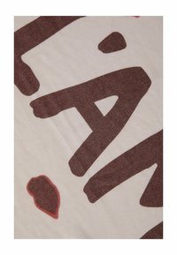 Tissu blanc cassé avec de grandes lettres brunes dessinées à la main et à l'aspect texturé, accompagnées d'un détail de tache d'encre rouge.