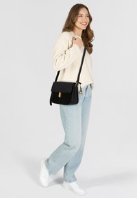 Bolso de hombro de cuero negro con tapa, detalle de broche dorado y correa ajustable. Combinado con vaqueros de un azul claro y un suéter crema.