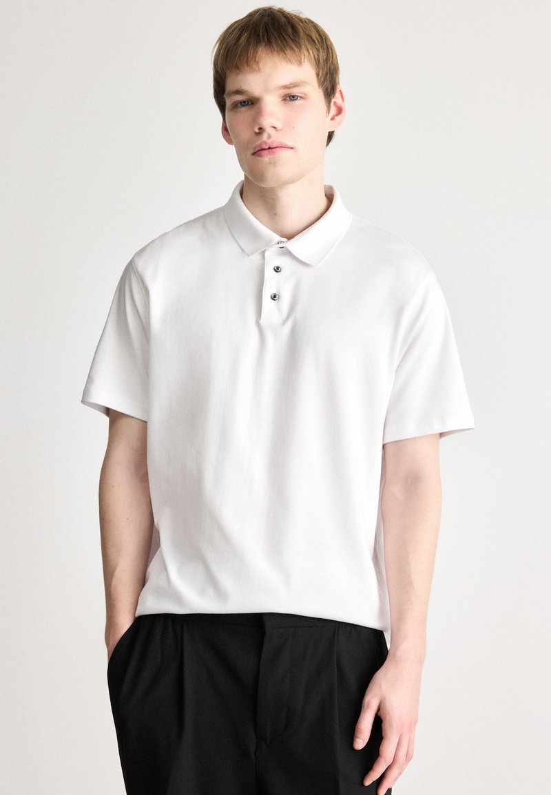 Polo-shirt en coton blanc avec un col à rabat, des manches courtes et une patte de boutonnage à trois boutons ; texture lisse et design minimaliste.