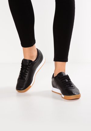 Czarne niskie trampki z białymi środkowymi podeszwami i gumowymi podeszwami noszone z czarnymi spodniami, stopy ustawione tak, że jedna jest lekko uniesiona.