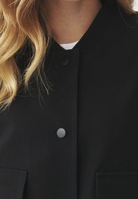 Černá bombarďáková bunda s kulatým výstřihem, předním zapínáním na knoflíky a dvěma náprsními kapsami. Hladká tkanina s matným povrchem.