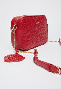 Bolso bandolera de imitación cuero acolchado rojo con una correa de cadena dorada y un charm en forma de rosa desmontable. Detalle del logo dorado en la parte frontal.