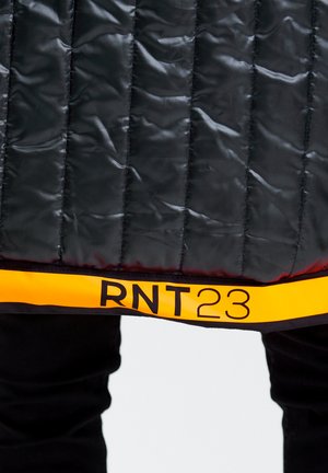 Zwarte gewatteerde jas met een glanzende textuur, voorzien van een oranje band aan de onderrand met "RNT23" in vetgedrukte zwarte letters.