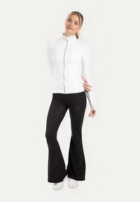 Giacca bianca con zip e collo alto, abbinata a pantaloni neri svasati. I materiali sono spandex o simili, creando una silhouette aderente.
