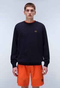 Granatowy sweter z okrągłym logo, okrągłym dekoltem, długimi rękawami i ribbrowymi mankietami, dopasowany do jasnopomarańczowych szortów. Gładka tekstura tkaniny.