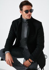 Schwarzer Wollmantel mit hohem Kragen, kombiniert mit einem grauen gerippten Rollkragenpullover. Die Person trägt runde Sonnenbrillen und dunkle Jeans und sitzt auf einem Drahtstuhl.