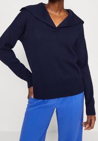Navy knit sweater with a wide collar and V-neck, featuring ribbed cuffs and hem. Paired with light blue trousers.