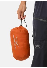 Oranje lichtgewicht opbergtasje met trekkoordsluiting en logo. Glad van structuur, ronde vorm, vastgehouden door een hand nabij donker kleding.