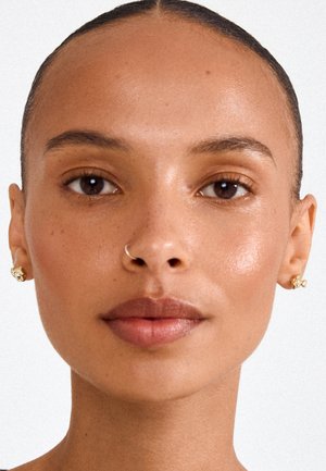 Junge Frau mit glatter Haut, trägt kleine goldene Ohrringe und einen silbernen Nasenring, schaut direkt in die Kamera vor einfachem Hintergrund.