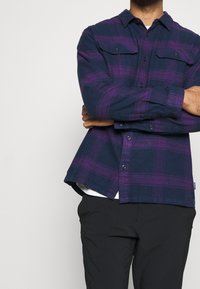 Nappitettava flanellipaita tummansinisellä pohjalla ja violettiruutuisella kuviolla. Siinä on kaksi rintataskua, kaulus ja hieman kaareva helma.