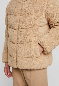 Beige fleece jas met een textuur die fluffy aanvoelt. Heeft horizontaal gestikte secties, een rits aan de voorkant en zijzakken.