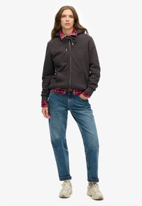 Sudadera gris con cremallera y capucha ajustable, combinada con una camisa de cuadros en rojo y negro, jeans azules y zapatillas de color claro.