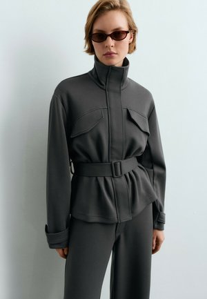 Vrouw draagt een grijze jas met ceintuur, klepzakken en hoge kraag, gecombineerd met bijpassende broek en donkere rechthoekige zonnebril.