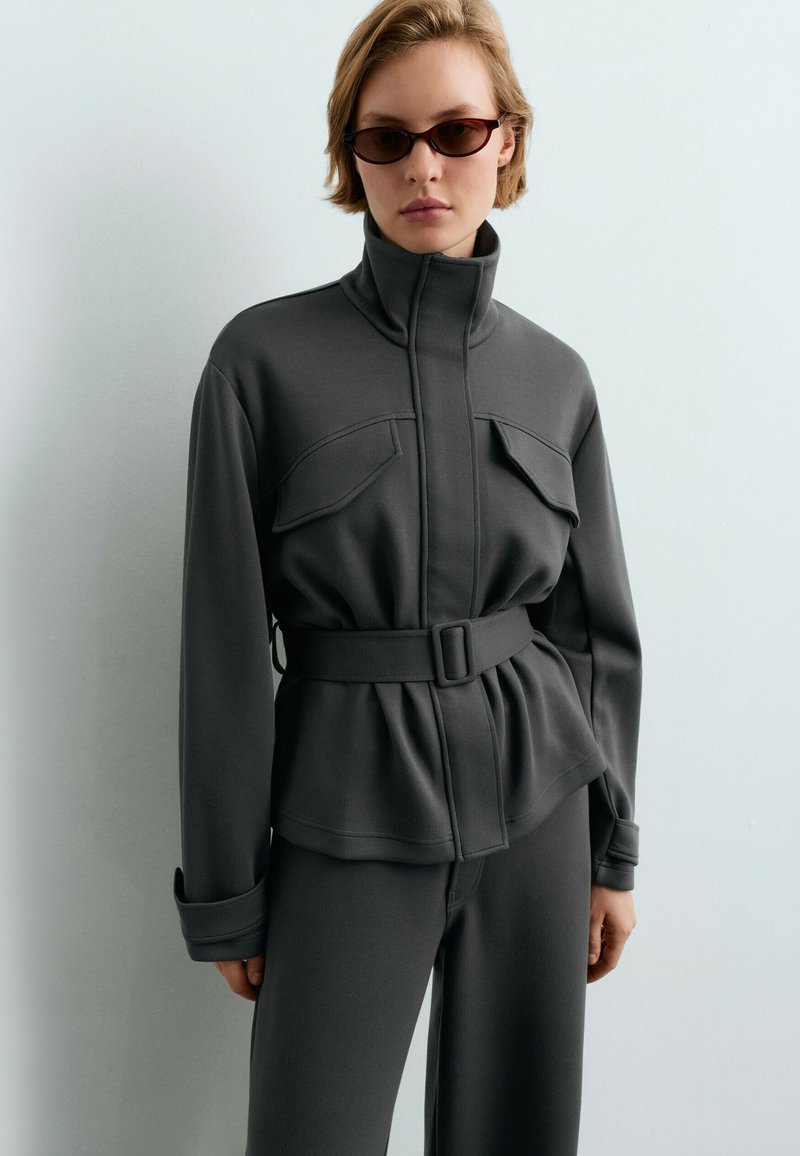 Femme portant une veste grise ceinturée avec des poches à rabat et un col montant, associée à un pantalon assorti et des lunettes de soleil rectangulaires foncées.