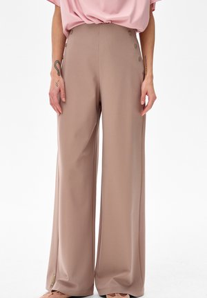 Beżowe szerokie spodnie z guzikami po bokach, noszone z luźnym różowym topem, ręce swobodnie opuszczone wzdłuż ciała na białym tle.