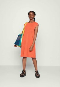 Vestido laranja com mangas curtas, corte solto, até aos joelhos. Acompanhado de uma mala multicolorida e sandálias pretas de plataforma. Design simples.