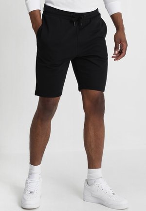 Mężczyzna w czarnych szortach ze sznurkiem, białych skarpetach i białych sneakersach Nike, stojący z rękami częściowo w kieszeniach na jednolitym tle.