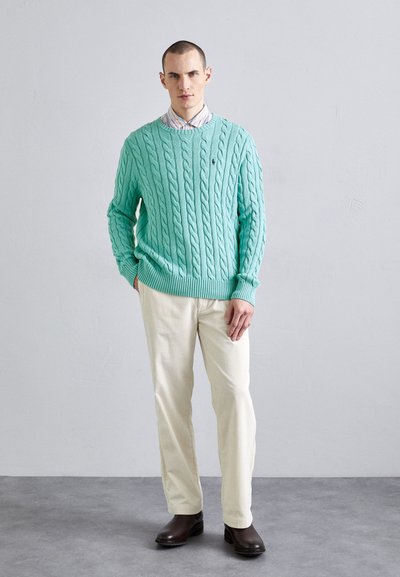 Pull en maille torsadée vert menthe, chemise à col rayée en dessous, pantalon en velours côtelé crème, bottes marron foncé. Le mannequin pose les mains dans les poches.