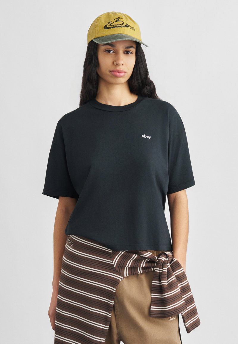 Schwarzes Baumwoll-T-Shirt mit kurzen Ärmeln und kleinem weißem "obey"-Logo, kombiniert mit gestreifter braun-weißer Hose und gelber bestickter Mütze.