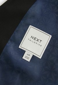 Tela de color azul marino con un patrón geométrico, con un borde negro. Etiqueta blanca que dice "NEXT TAILORING", talla 40R, con información de cuidado debajo.