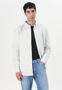 Chemise boutonnée rayée vert clair en coton, avec un col classique, portant un t-shirt noir en dessous et un jean en denim.