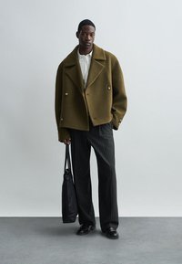 Olijfgroen wollen dubbelzijdig jasje, witte overhemd, gestreepte broek, zwarte tas en zwarte schoenen, tegen een neutrale achtergrond.