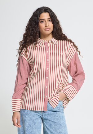 Femme aux longs cheveux bouclés portant une chemise à rayures crème et rouges et un jean bleu clair, debout avec une main dans la poche.