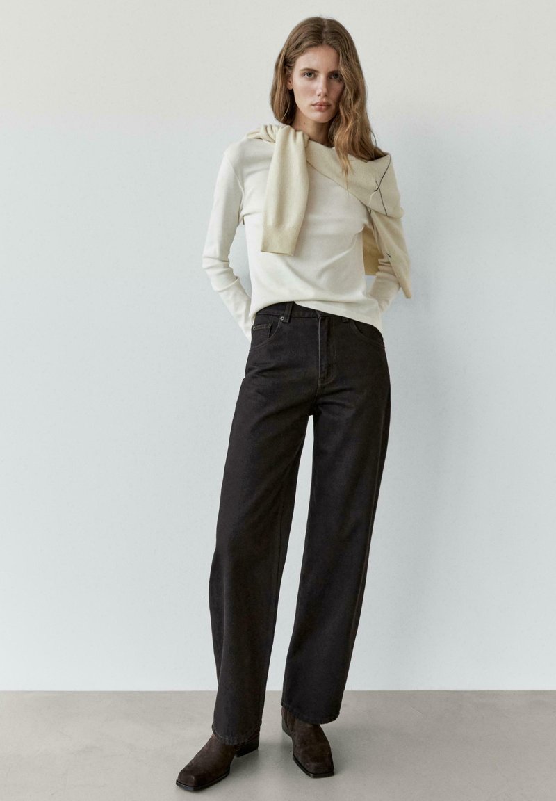 Beige top met lange mouwen en een licht gebreide sjaal om de hals gedrapeerd, gecombineerd met zwarte jeans met wijde pijpen en donkerbruine enkellaarsjes.