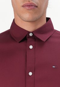 Bordeaux blouse met een button-down kraag, gemaakt van een gladde stof. Voorzien van witte knopen en een klein geborduurd logo in rood, wit en blauw.