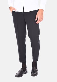 Pantaloni neri su misura con una vestibilità aderente, caratterizzati da una consistenza liscia e un orlo cuff. Indossati con mocassini neri e calze scure.