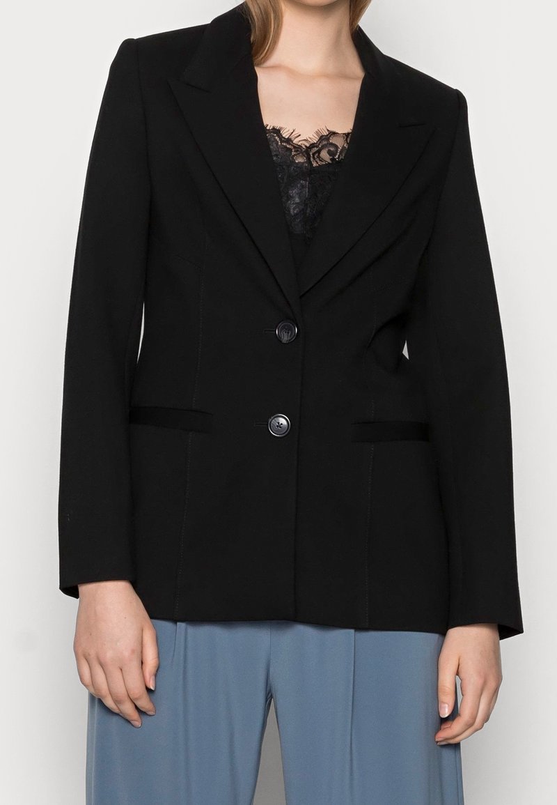 Femme portant un blazer noir ajusté à deux boutons sur un top en dentelle noire et un pantalon bleu clair ample.