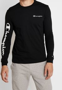 Schwarzes langärmliges Baumwollshirt mit weißem "Champion"-Logo auf der Brust und entlang des linken Ärmels.