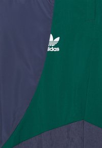Zelená a námořnicky modrá mikina s rozdělenými panely a texturovaným povrchem. Na zelené části je bílý logo Adidas. Detail zipu není viditelný.
