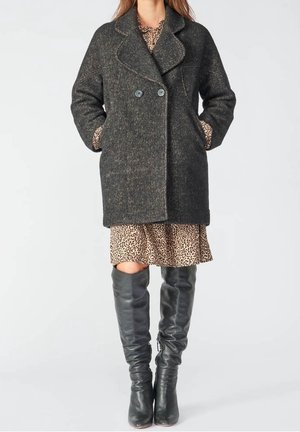 Frau trägt einen dunkelgrauen zweireihigen Mantel, ein Kleid mit Leopardenmuster und schwarze kniehohe Lederstiefel, steht mit den Händen in den Manteltaschen.