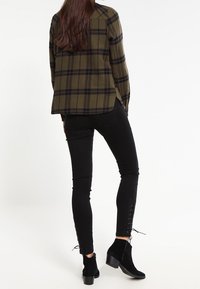 Grünes und schwarzes kariertes Hemd mit seitlichen Schlitzen, kombiniert mit schwarzen Skinny-Jeans mit Schnürdetails an den Knöcheln und schwarzen Stiefeletten.