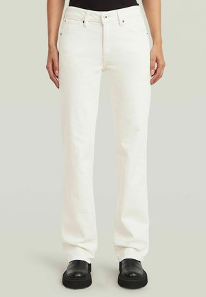Femme portant un jean blanc droit et des chaussures compensées noires, debout devant un fond clair uni.