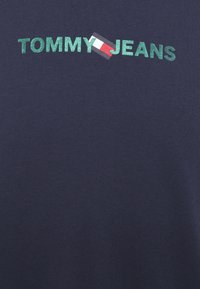 Mörkblå bomullströja med "TOMMY JEANS" i grön glitter och en logotypikon i rött, vitt och blått på bröstet.